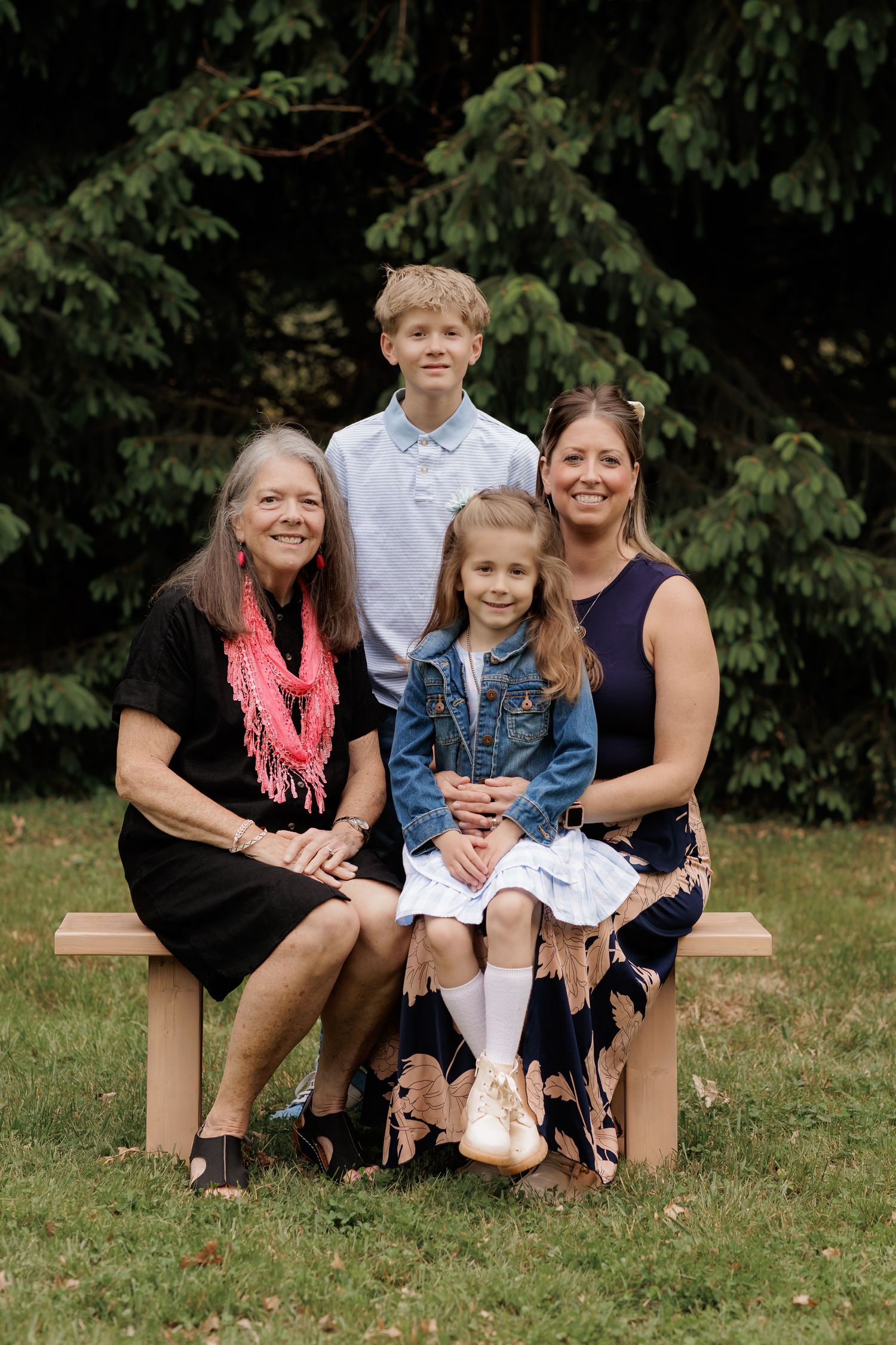 Family portrait of two women and two children sitting on a wooden bench outdoors with lush green trees in background.