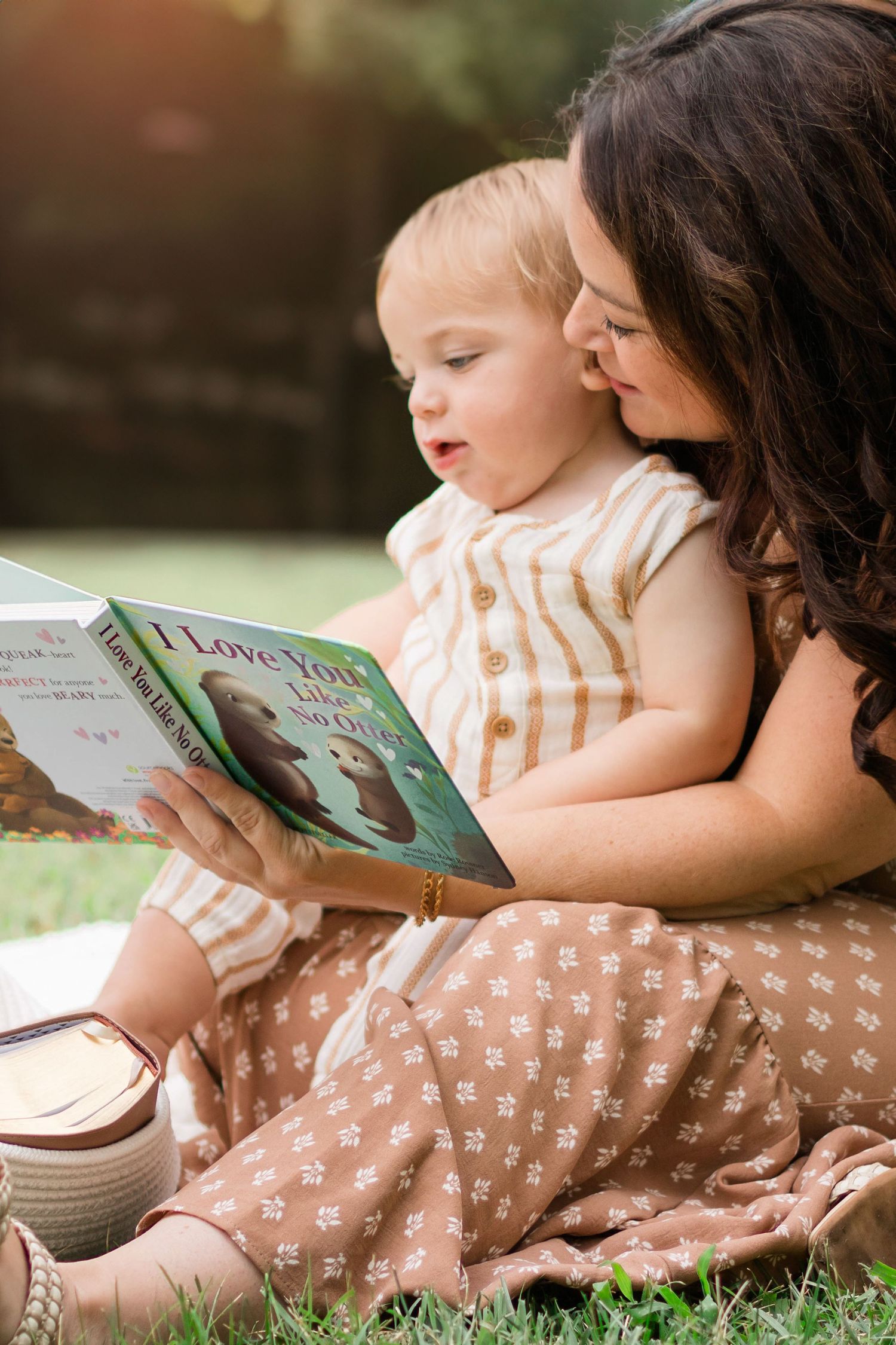 Mother and baby sitting on grass reading 'I Love You to the Moon and Back' children's book together outdoors.