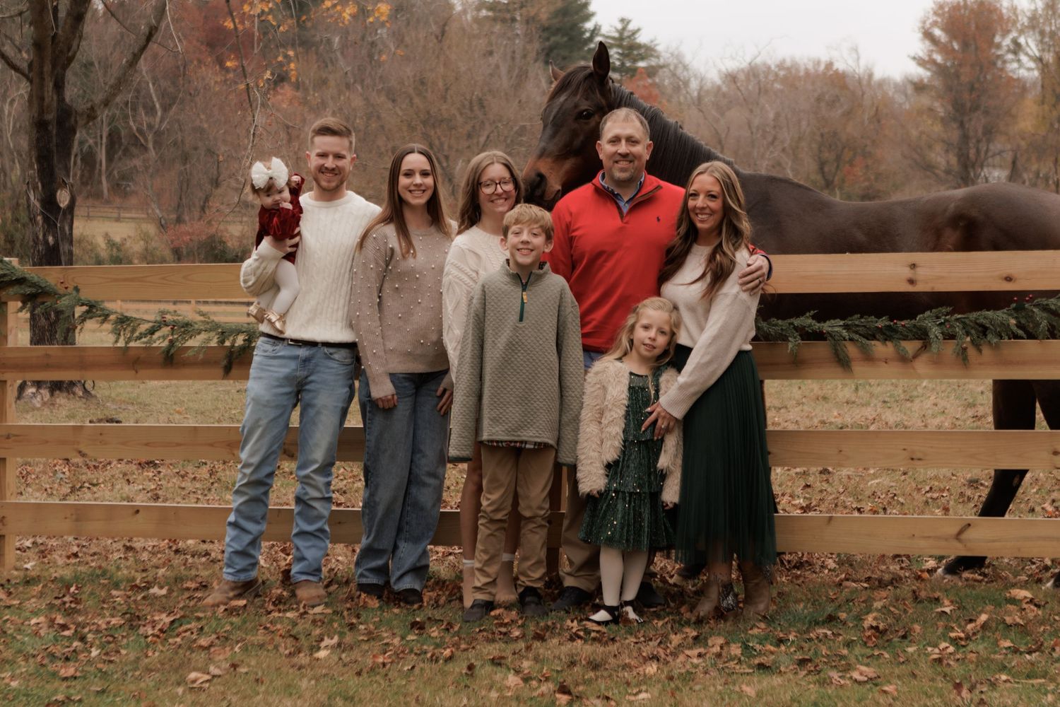 Family of seven posing with a horse by a wooden fence during autumn, surrounded by colorful fallen leaves.