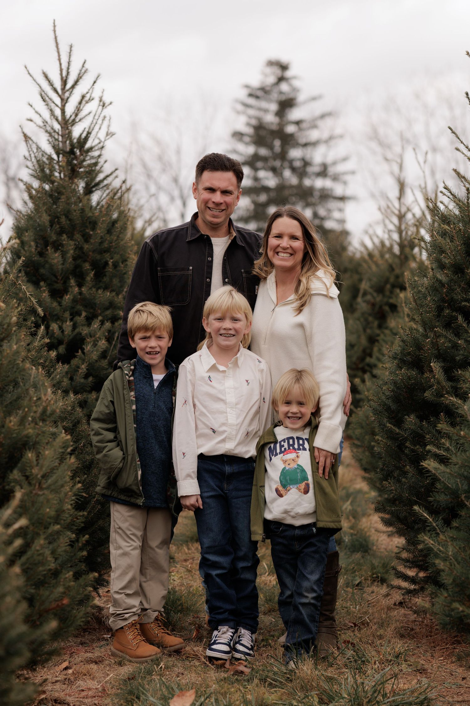 Family of five posing among evergreen trees at a Christmas tree farm, parents with three young blonde children smiling.