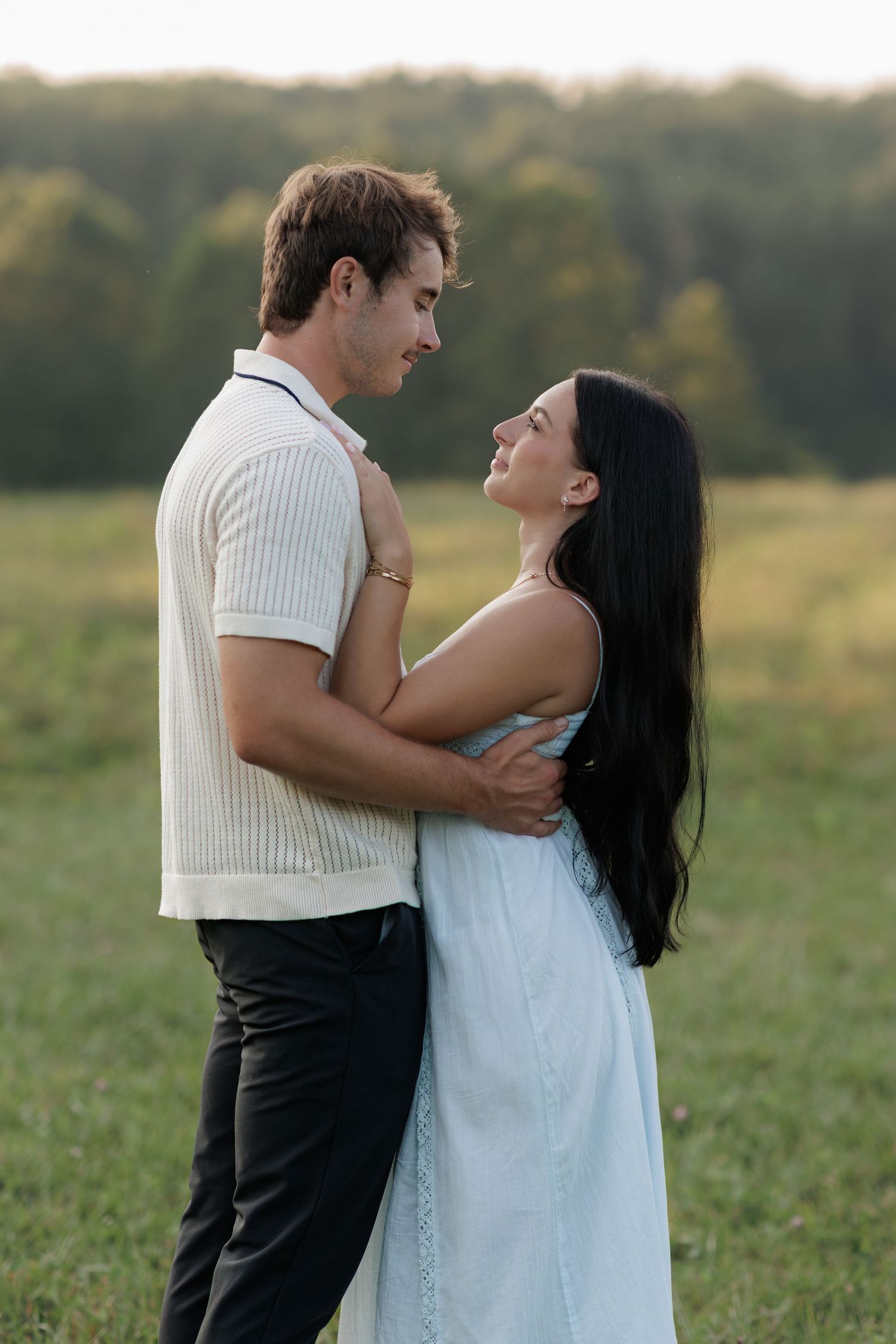 A couple embraces in a green field, the woman in a light blue dress gazing up at the man in a white shirt.