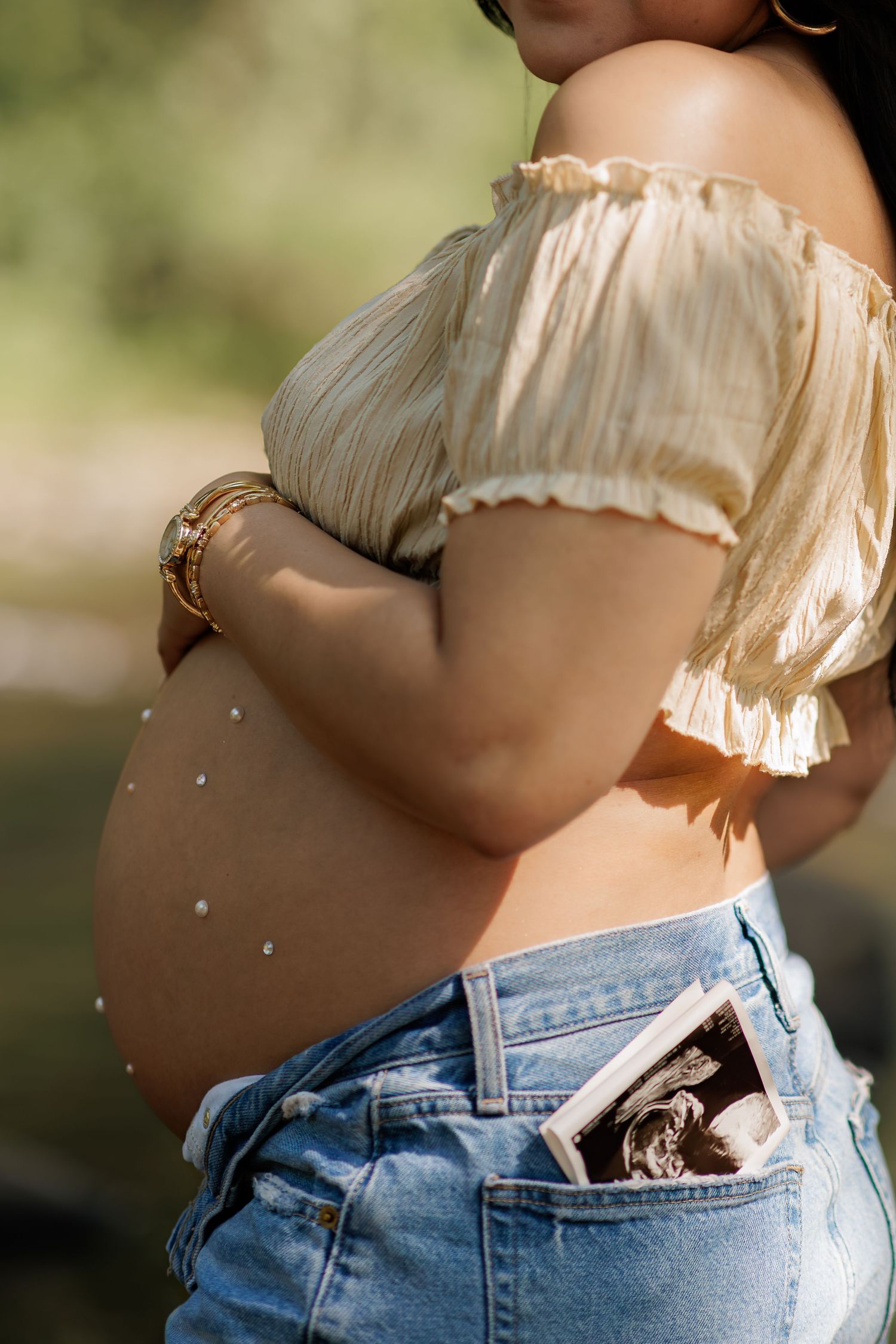 Pregnant woman in a cream off-shoulder top and denim jeans cradling her pearl-decorated baby bump with an ultrasound photo in her pocket.