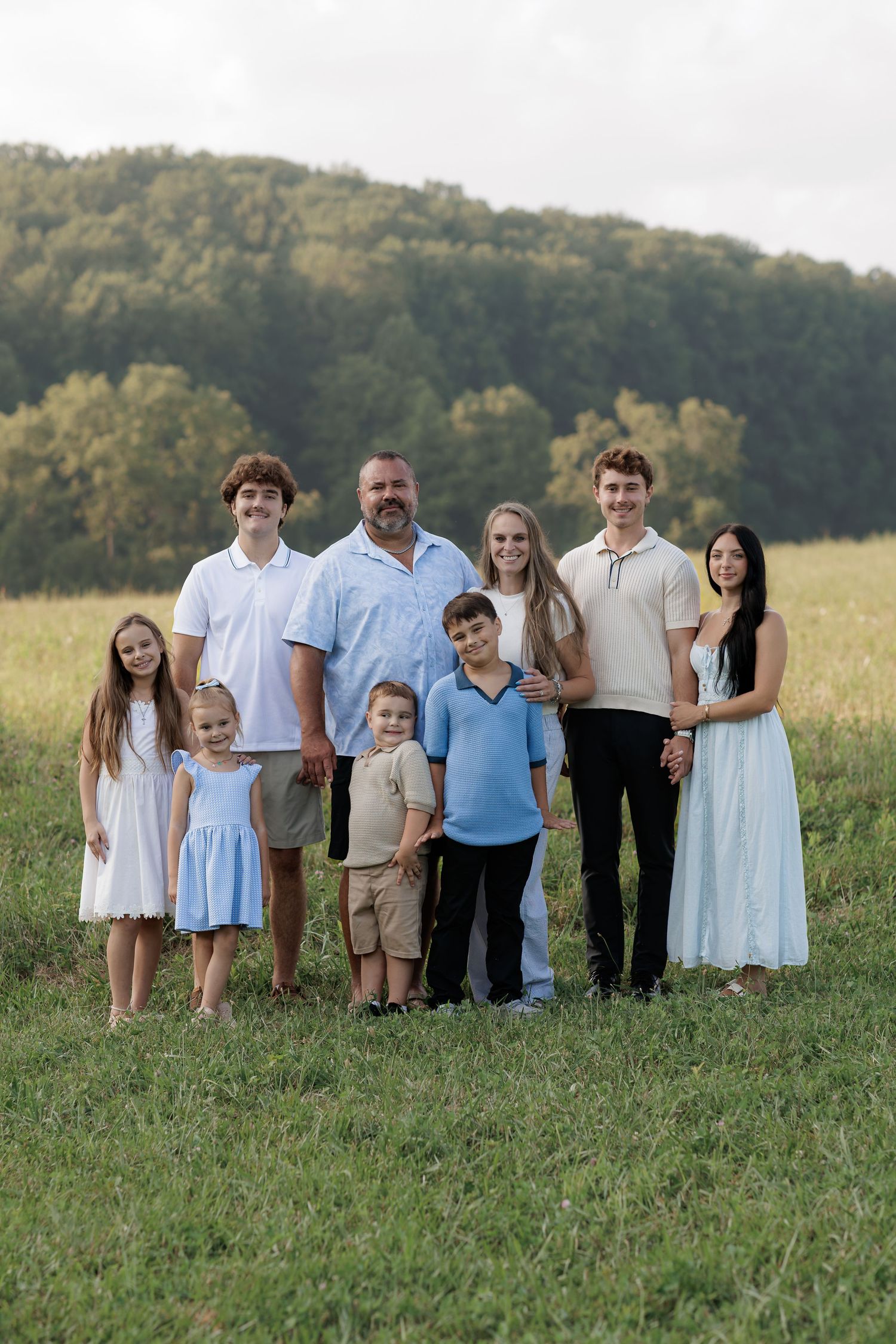 Large family portrait with parents and six children posing outdoors in a green field with wooded hills behind them.