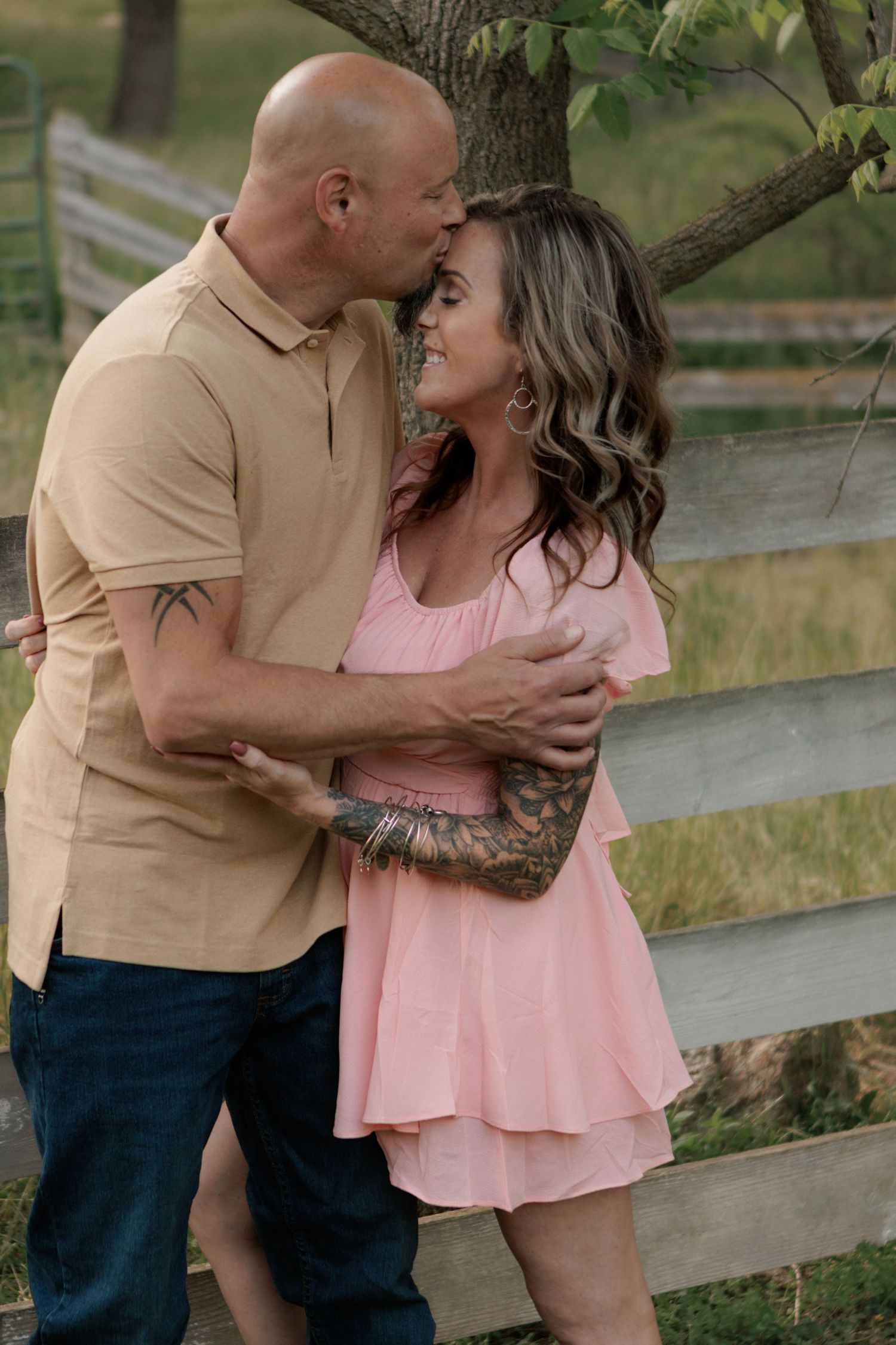 A bald man with tattoos kisses a woman in a pink dress near a wooden fence outdoors.