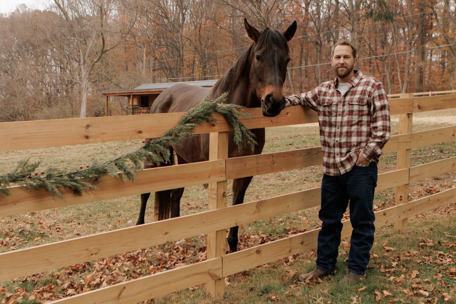 Man in plaid shirt stands beside a dark brown horse at a wooden fence decorated with evergreen garland in autumn.