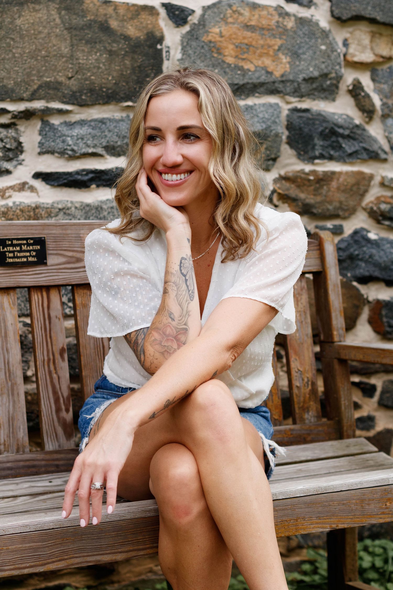 A woman in a white blouse sits on a wooden bench against a stone wall background.