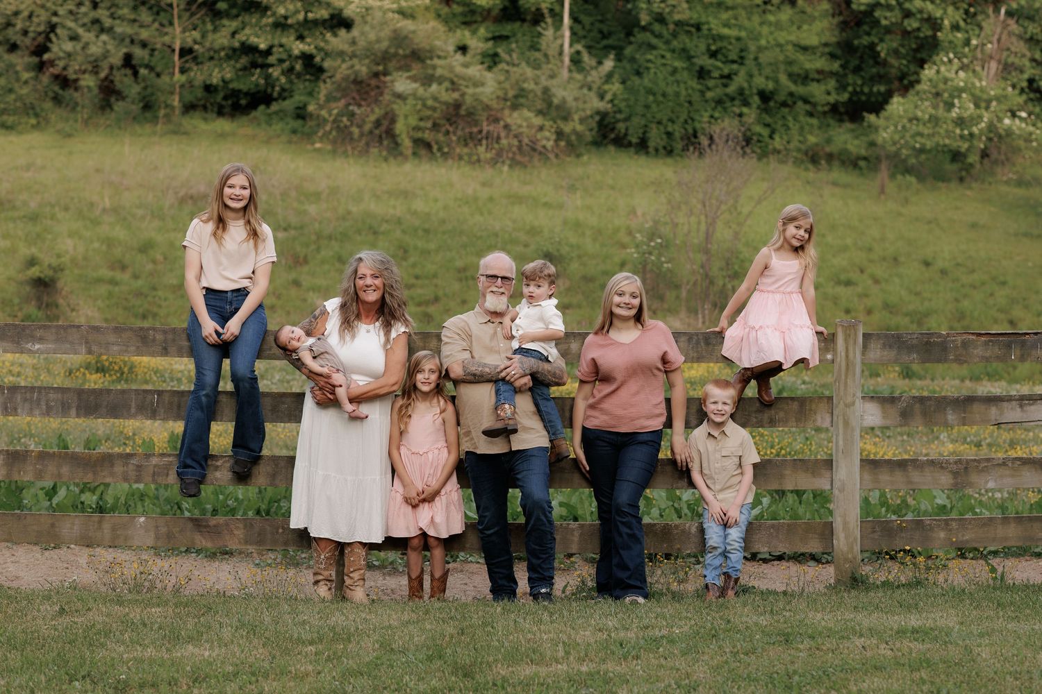 Multi-generational family portrait outdoors near wooden fence on green hillside, featuring grandparents with children and grandchildren.