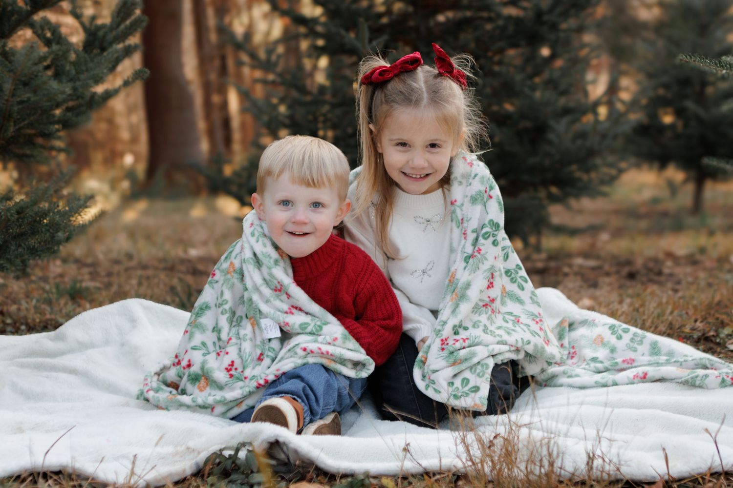 Two young children sharing a floral blanket outdoors among pine trees for a festive holiday photo session.