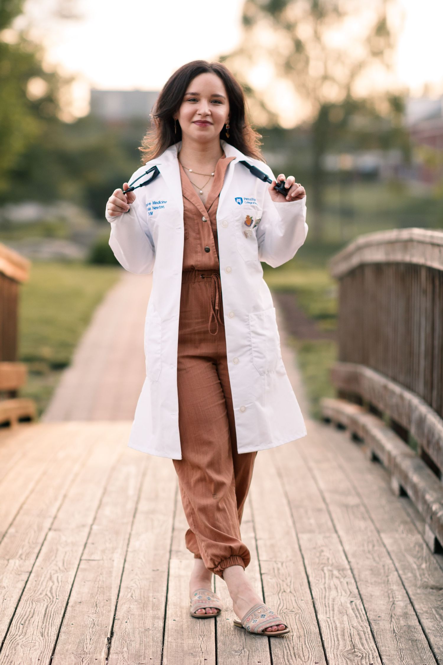Person in brown jumpsuit and white coat walks confidently across wooden bridge at sunset.