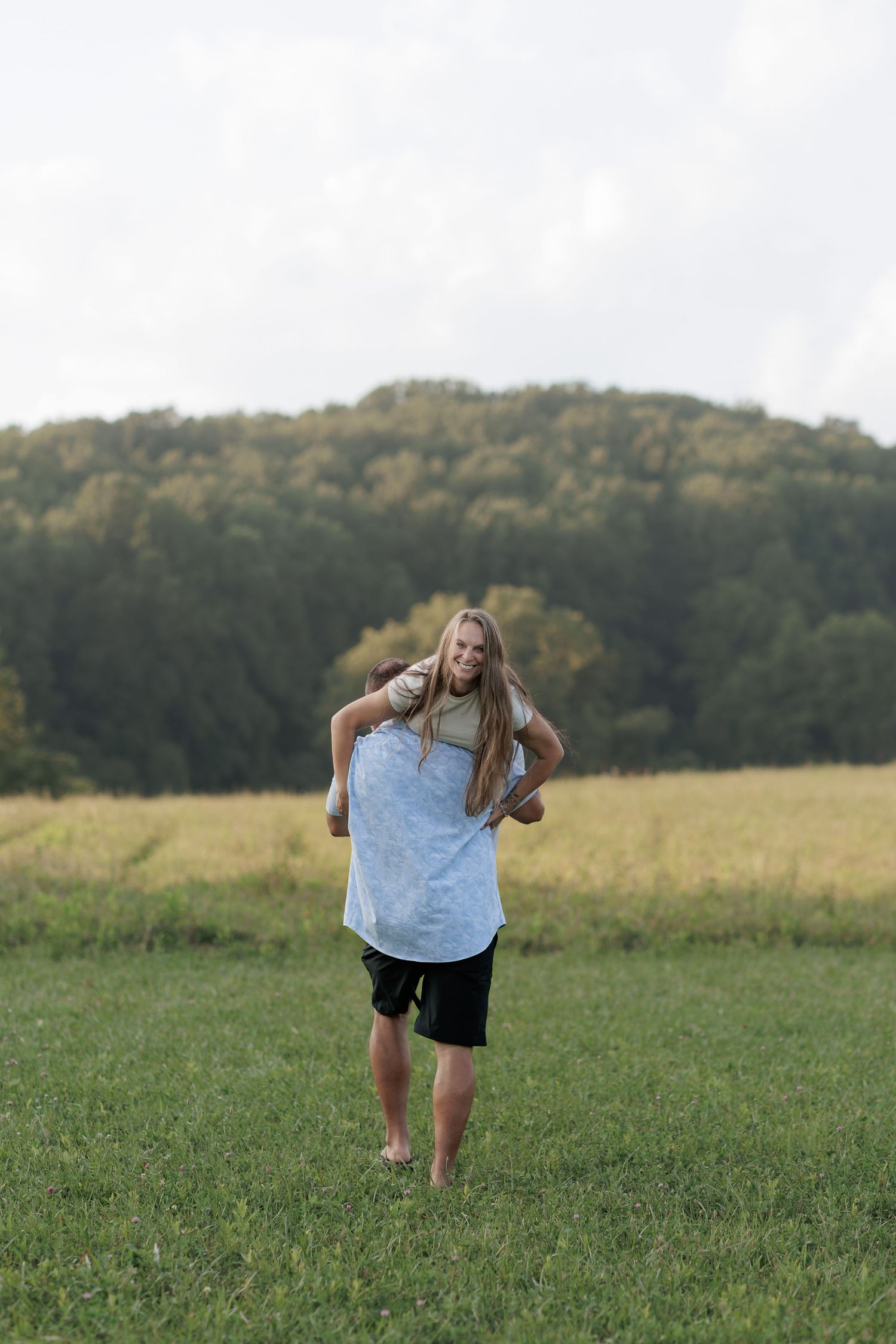 A man giving a woman a piggyback ride in a green field with trees in the background.