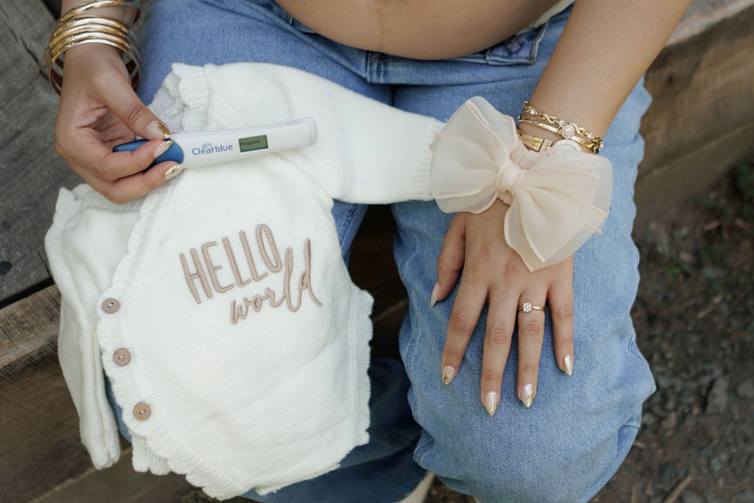 Woman in jeans holding a pregnancy test beside a white baby cardigan embroidered with Hello World and a cream bow.