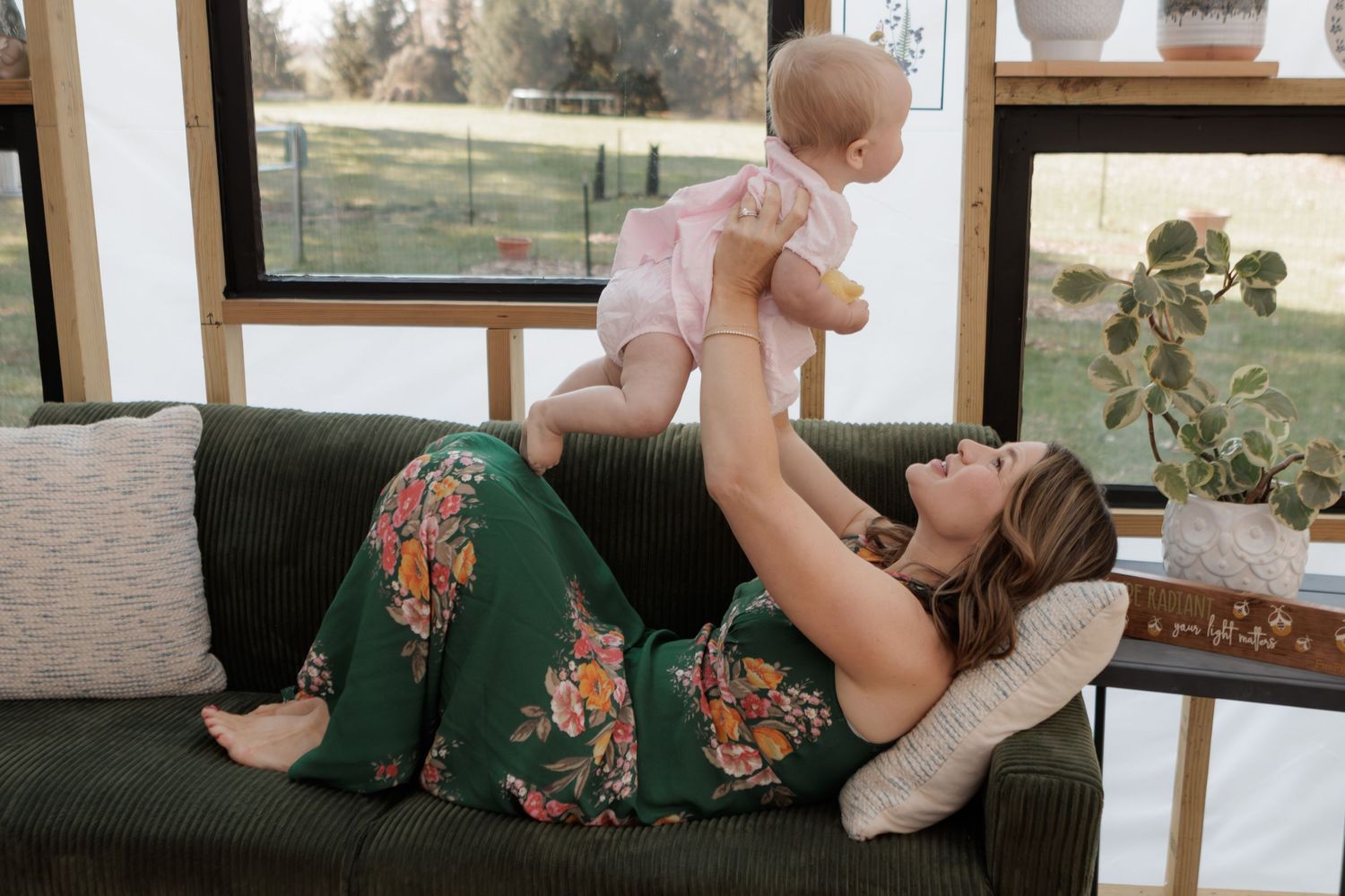 A mother lies on a green floral sofa, joyfully lifting her baby girl in a pink onesie in a bright sunlit room.