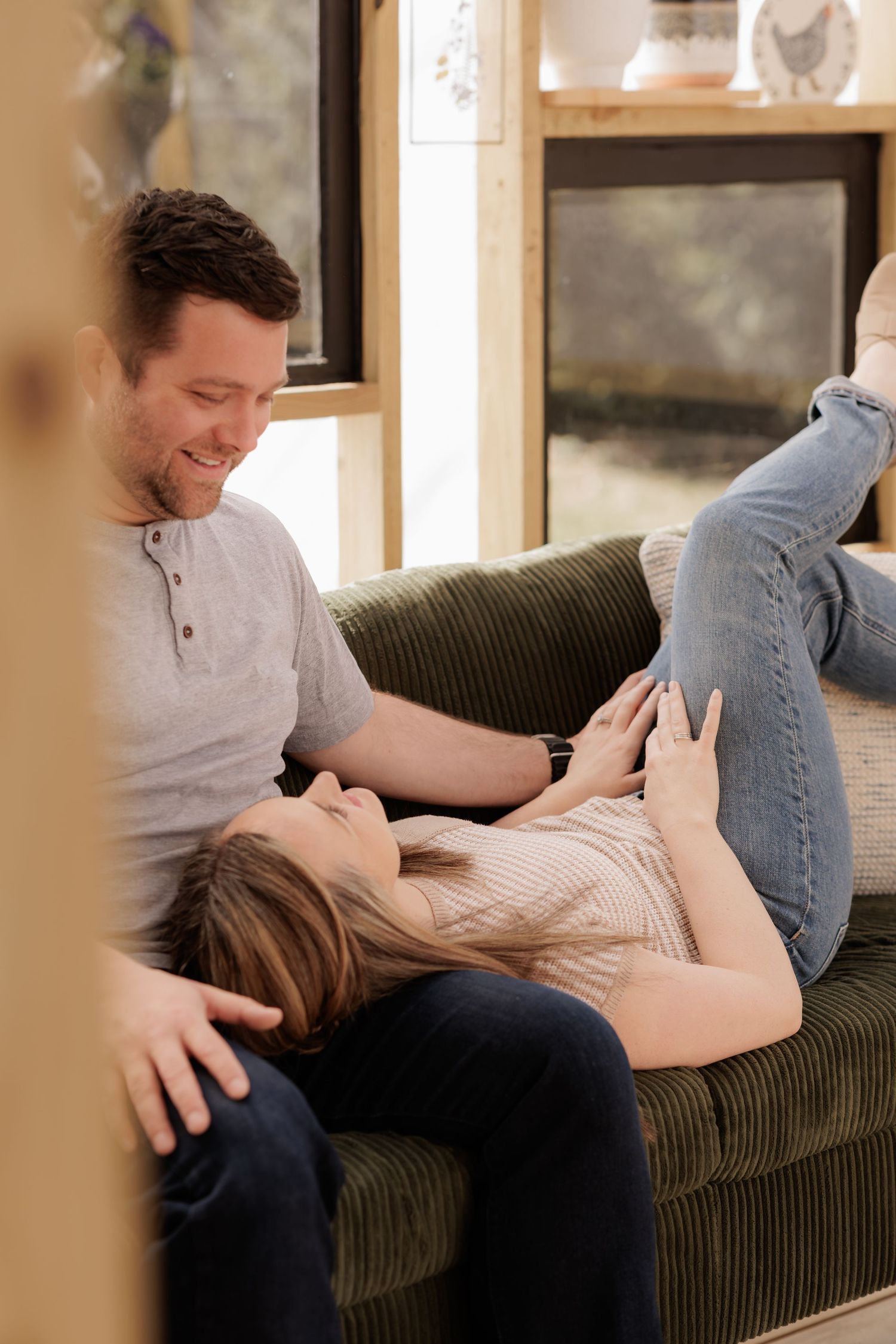 A couple relaxes on a green sofa, woman lying with head in mans lap while he smiles down at her.