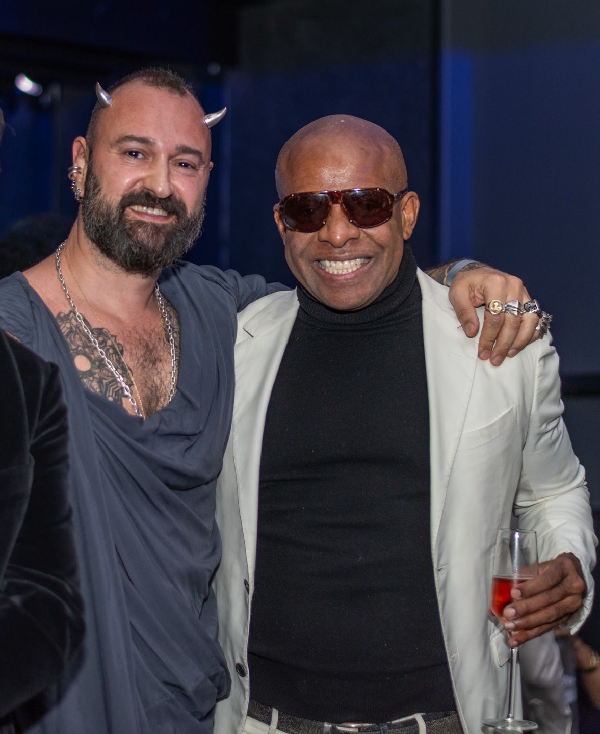 Two men smiling together at event, one with tattoos and devil horns, one in white blazer.