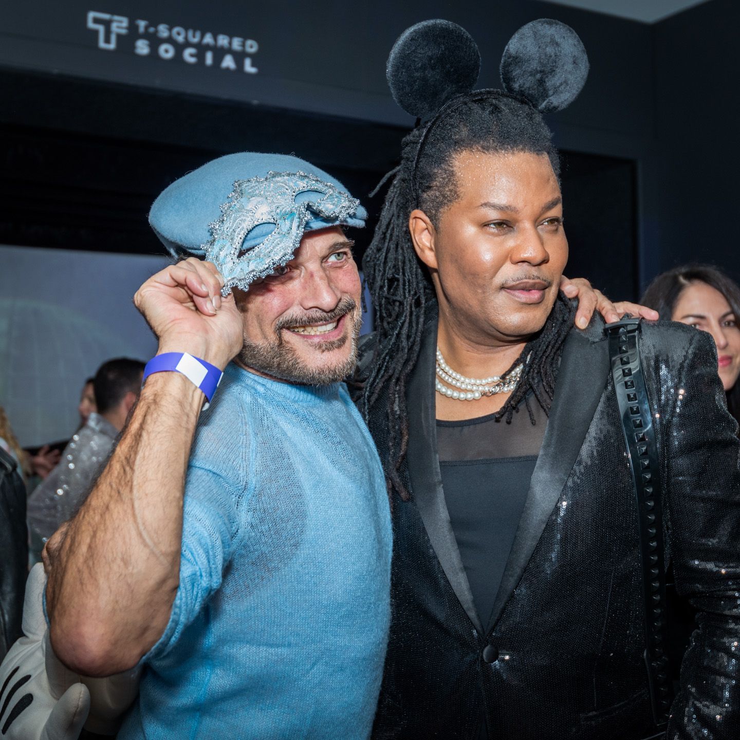 Two people in festive costumes pose at T-Squared Social event, one wearing a blue masquerade mask.