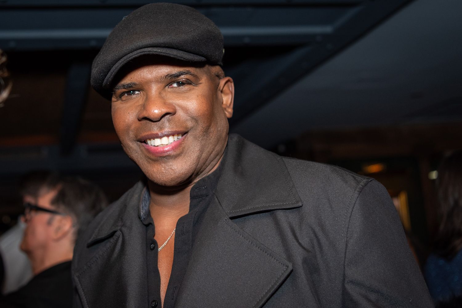 A smiling man wearing a black cap and jacket poses at an indoor social event.