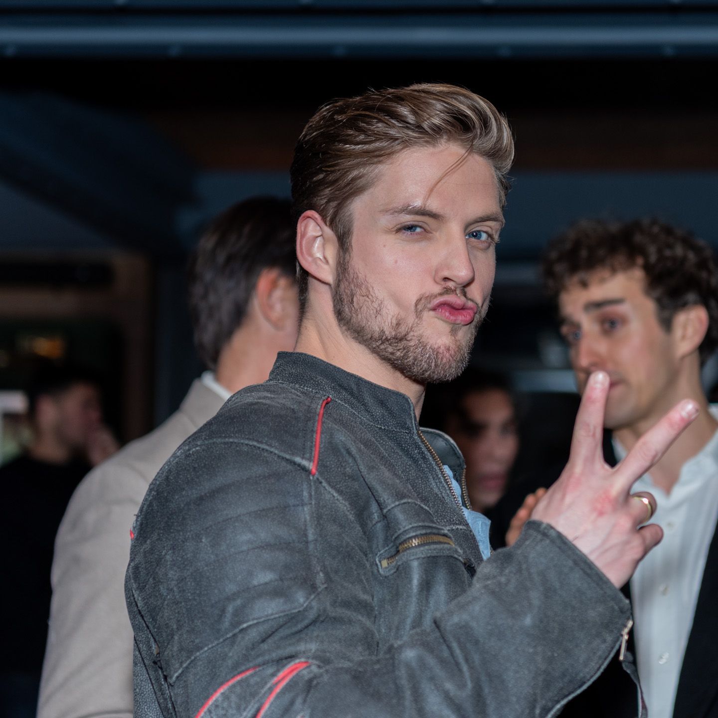 Blonde man in gray leather jacket making peace sign at nightclub event.