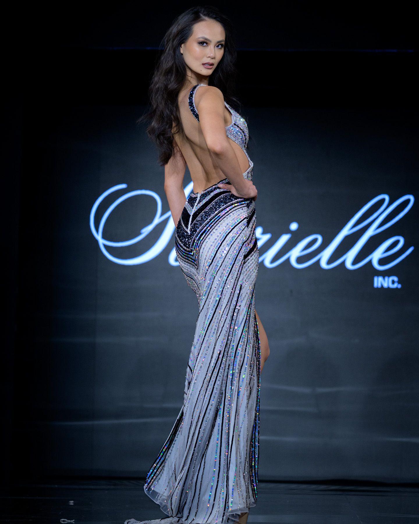 Model showcases backless striped sequin evening gown on dark fashion runway.