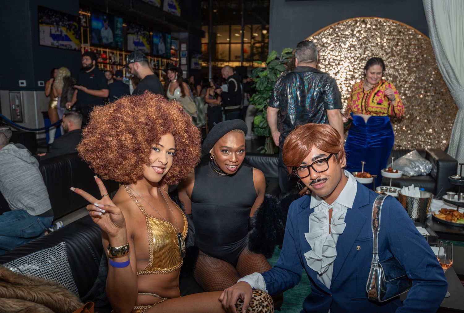 Three guests in 70s-inspired costumes posing at a themed nightclub party with gold disco decor.