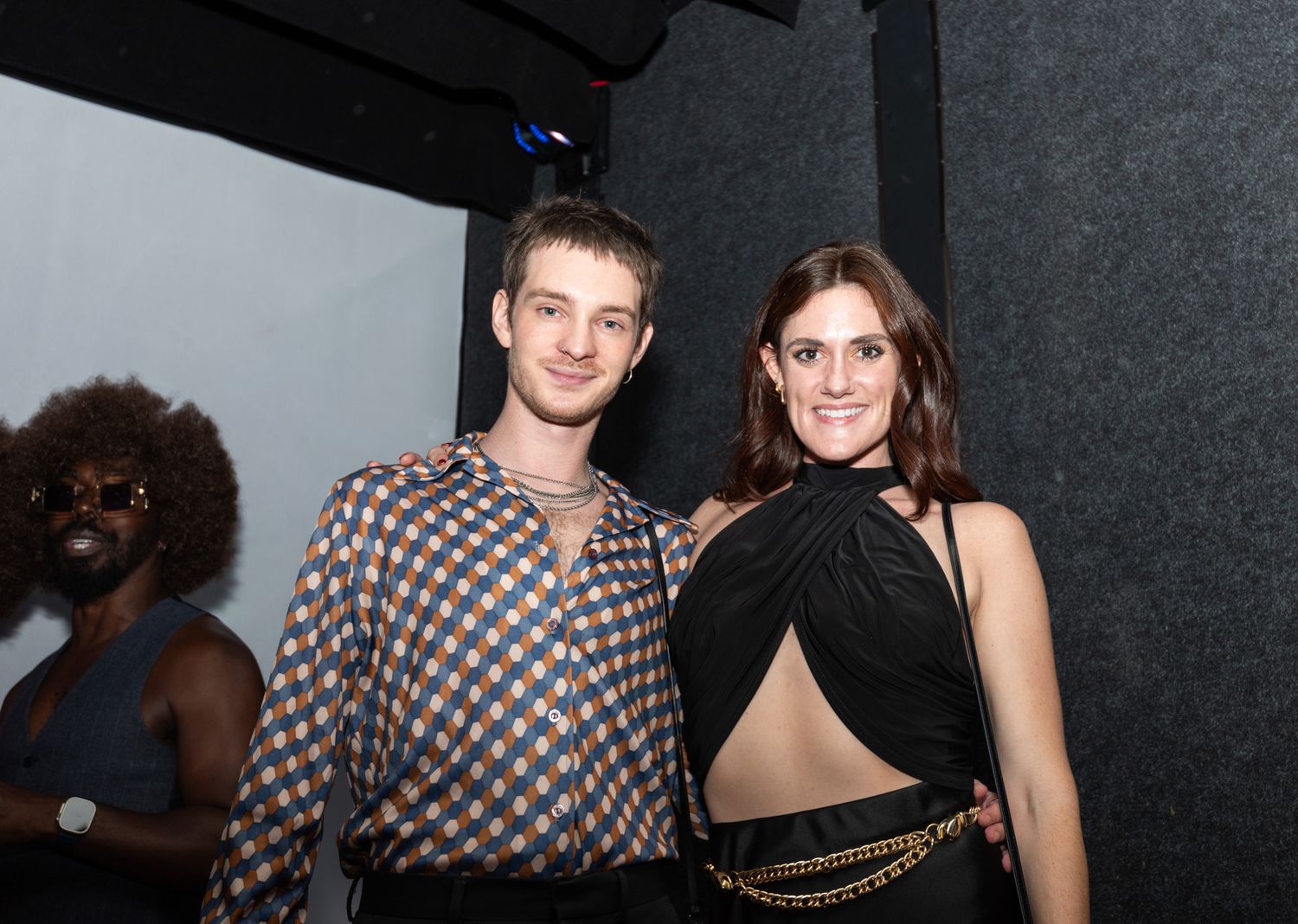 Young couple poses at an indoor evening event, man in patterned shirt and woman in black halter dress.