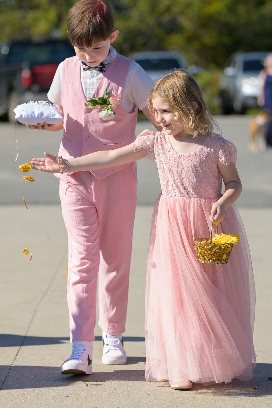 Ring bearer and flower girl in pink outfits scatter petals outdoors.