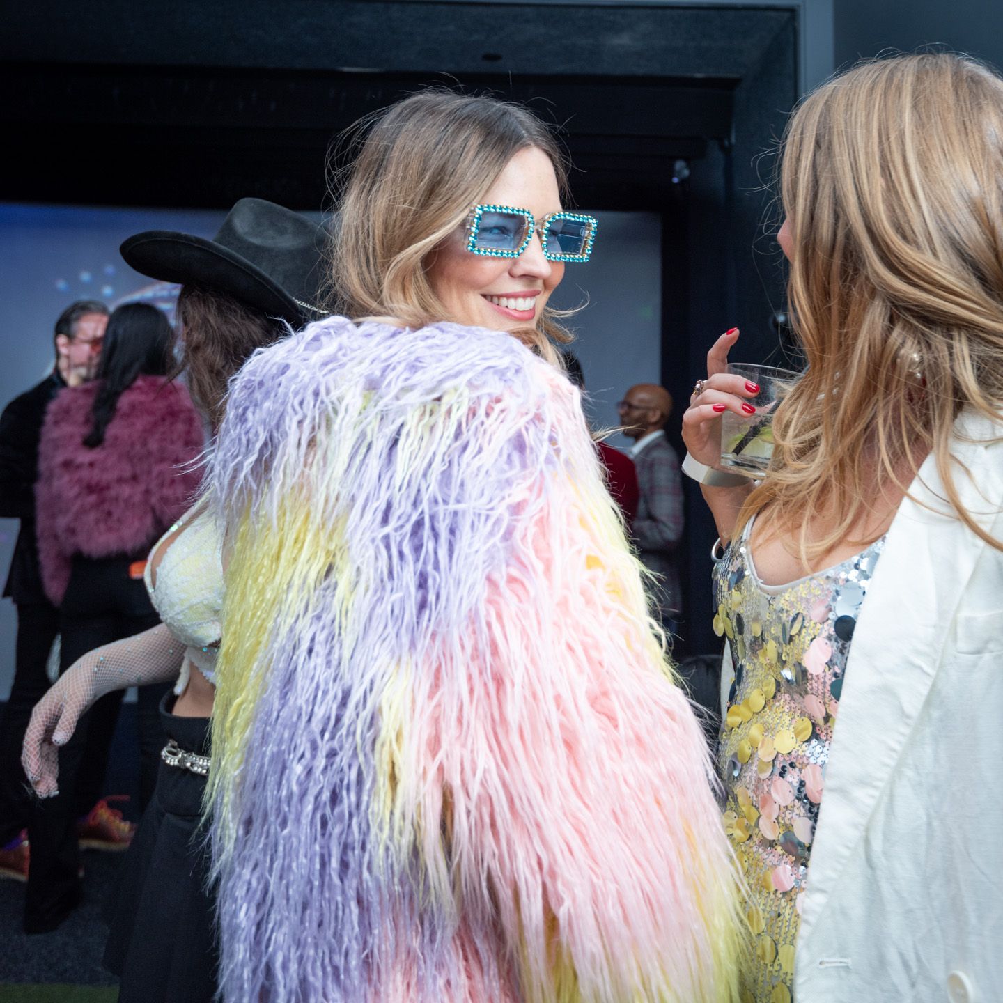 Woman in pastel rainbow shaggy fur coat and teal sunglasses at outdoor fashion event.