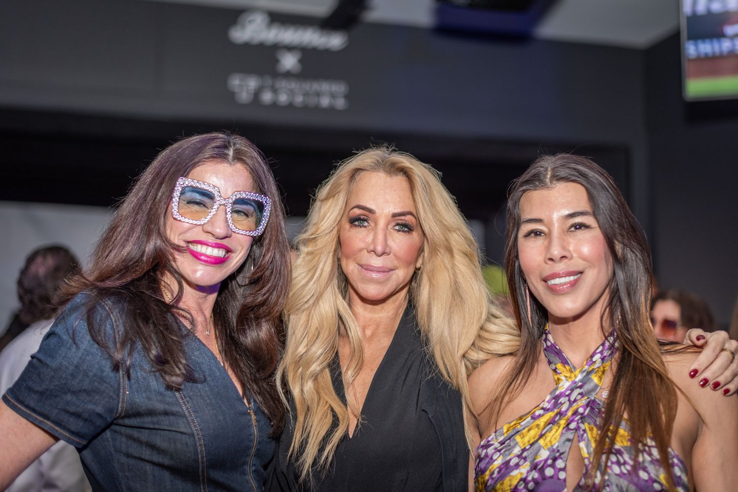 Three women smiling together at a T-Squared Social event, one wearing decorative glasses.
