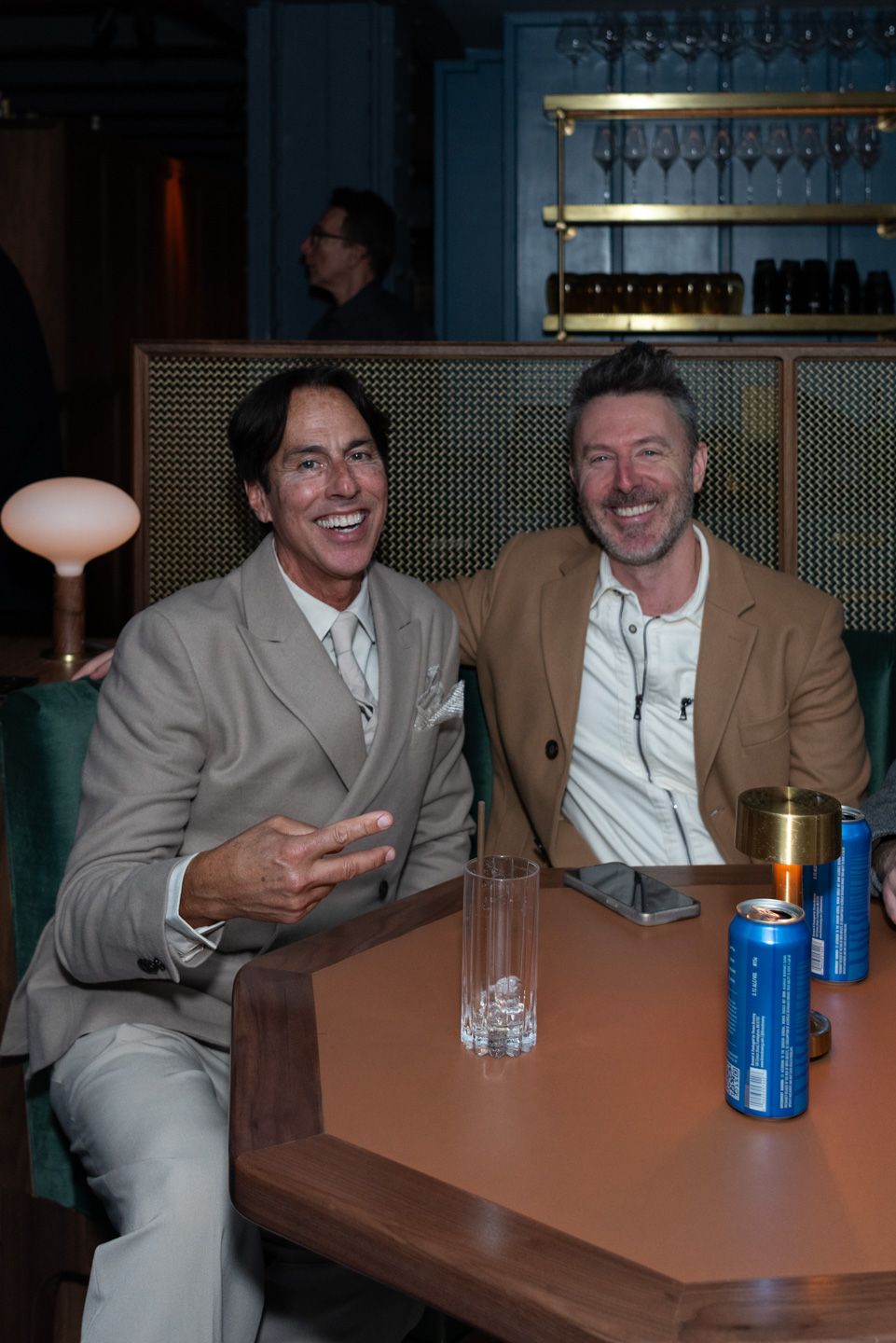 Two men laugh together at a table with beer cans in a dimly lit bar.