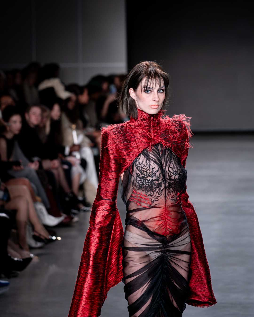 Model walks runway in dramatic red feathered coat over sheer black bodysuit at NYFW.