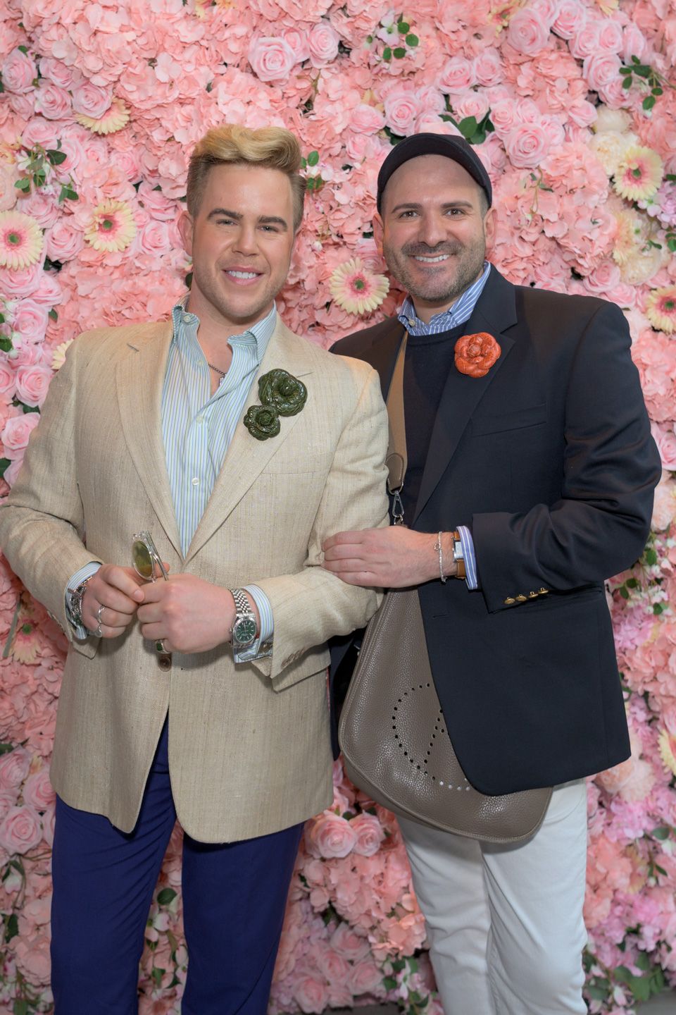 Two men in blazers with floral boutonnieres pose before a pink flower wall backdrop.