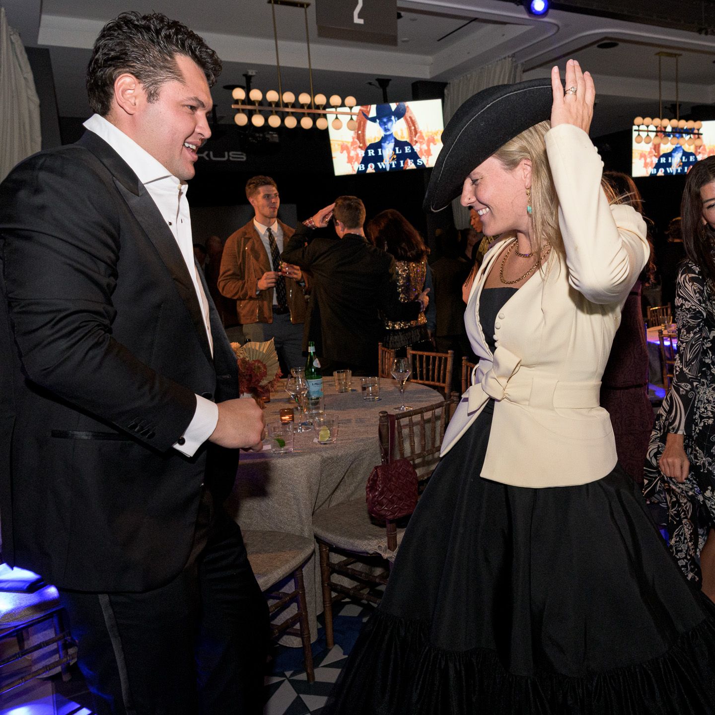 Man in tuxedo dances with woman wearing large black hat at gala.