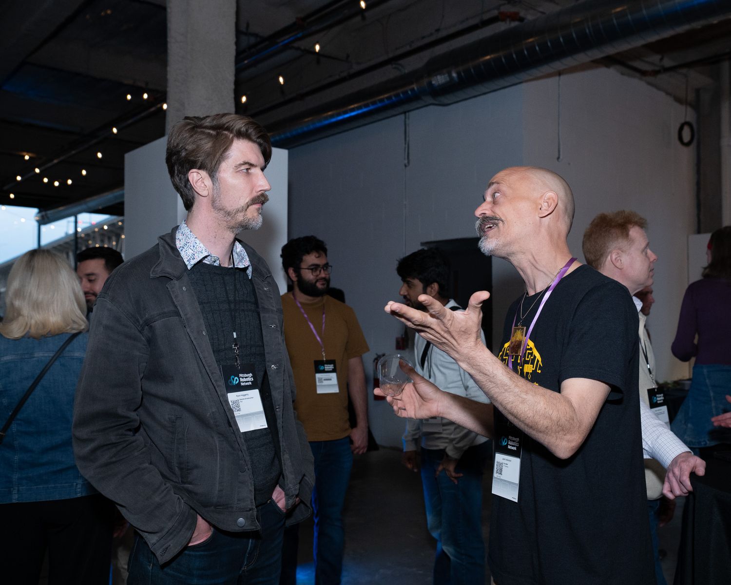 Two men with lanyards having an animated conversation at an indoor networking event.