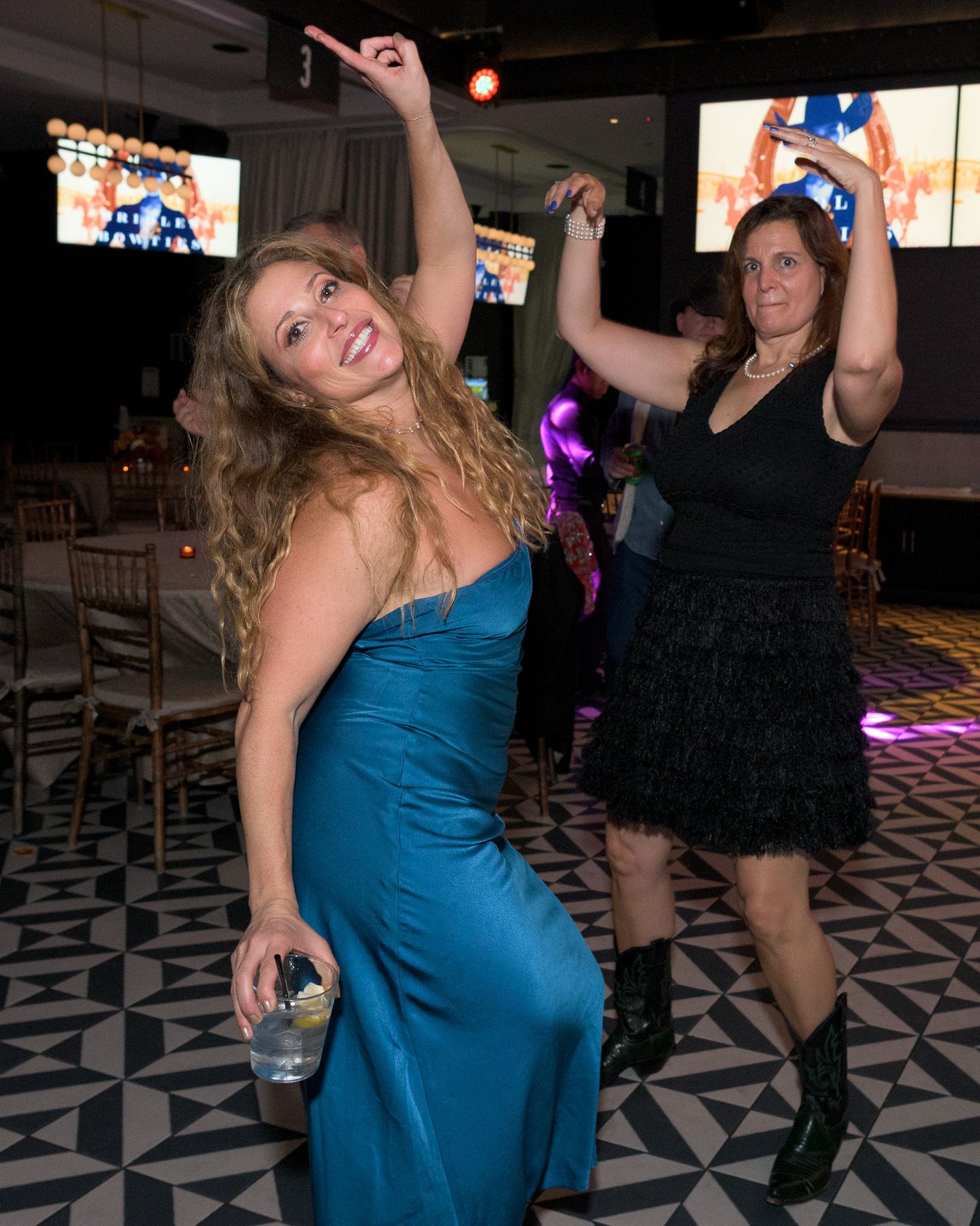 Two women dance energetically on a black and white patterned floor at a party.