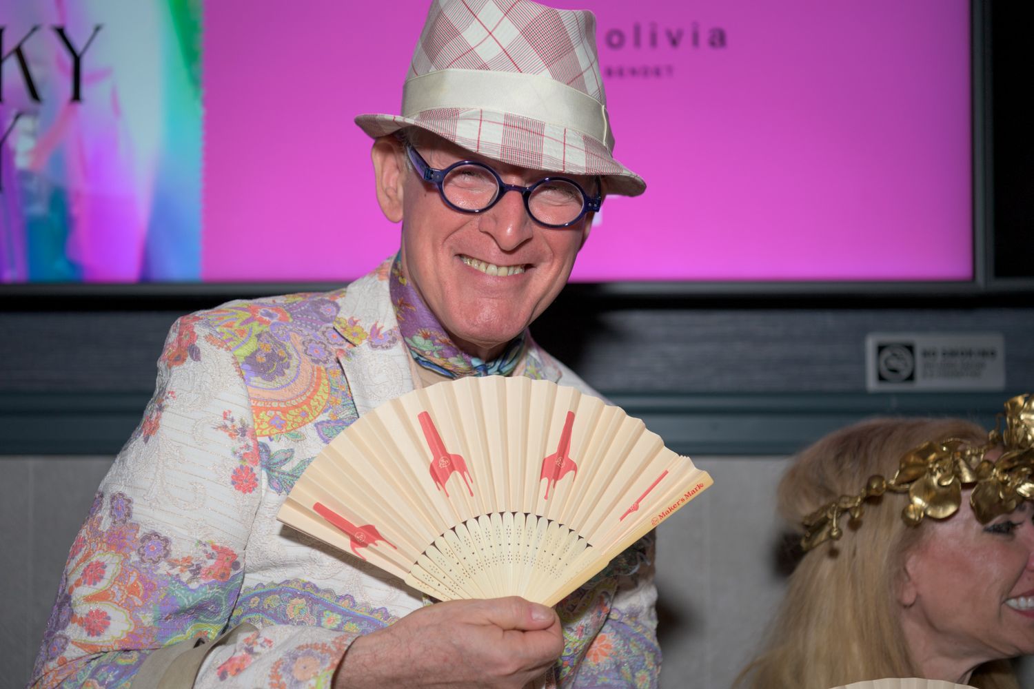 Elegantly dressed man in floral jacket and hat holds decorative fan at festive event.