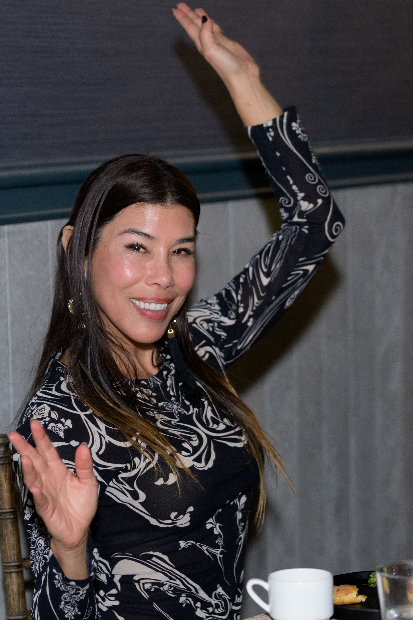 Woman in black and white patterned dress raises hand and smiles at a formal charity dinner event.