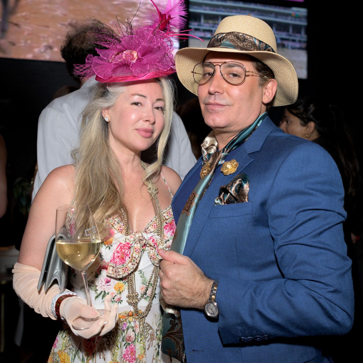 Couple posing at party, woman in floral dress with pink feathered hat, man in blue suit with panama hat.