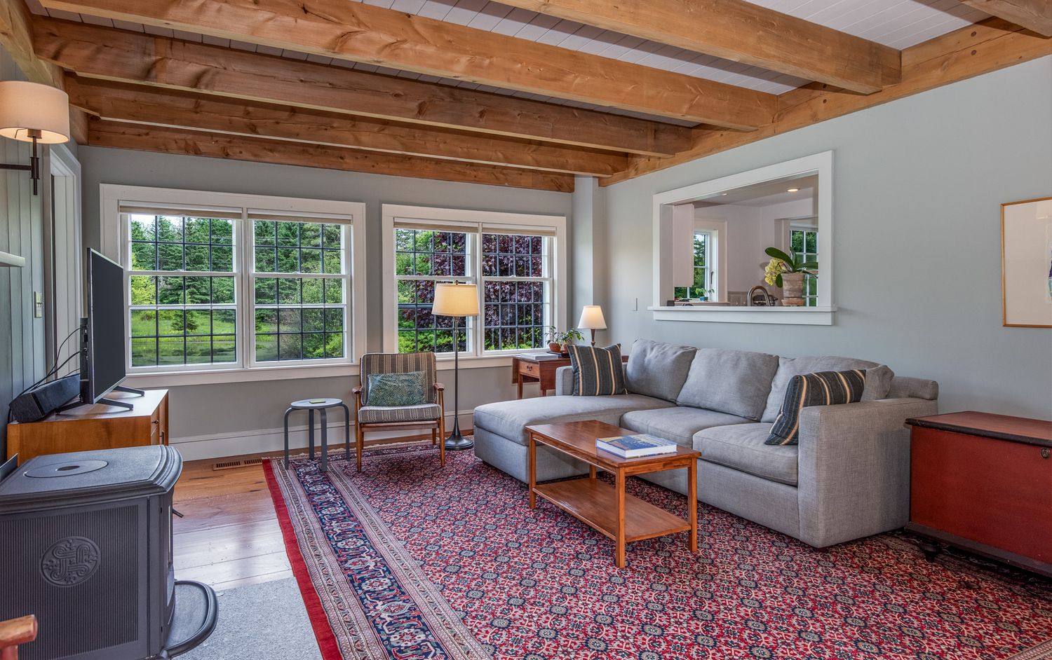 Bright living room with exposed wood beam ceiling, gray sofa, Persian rug, and large garden-view windows.