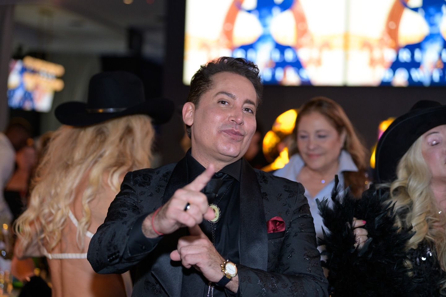 Stylish man in black sequin blazer points at camera at an upscale charity gala event.