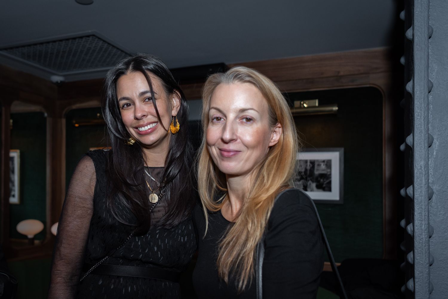 Two women posing together for a photo at a dimly lit indoor social event.