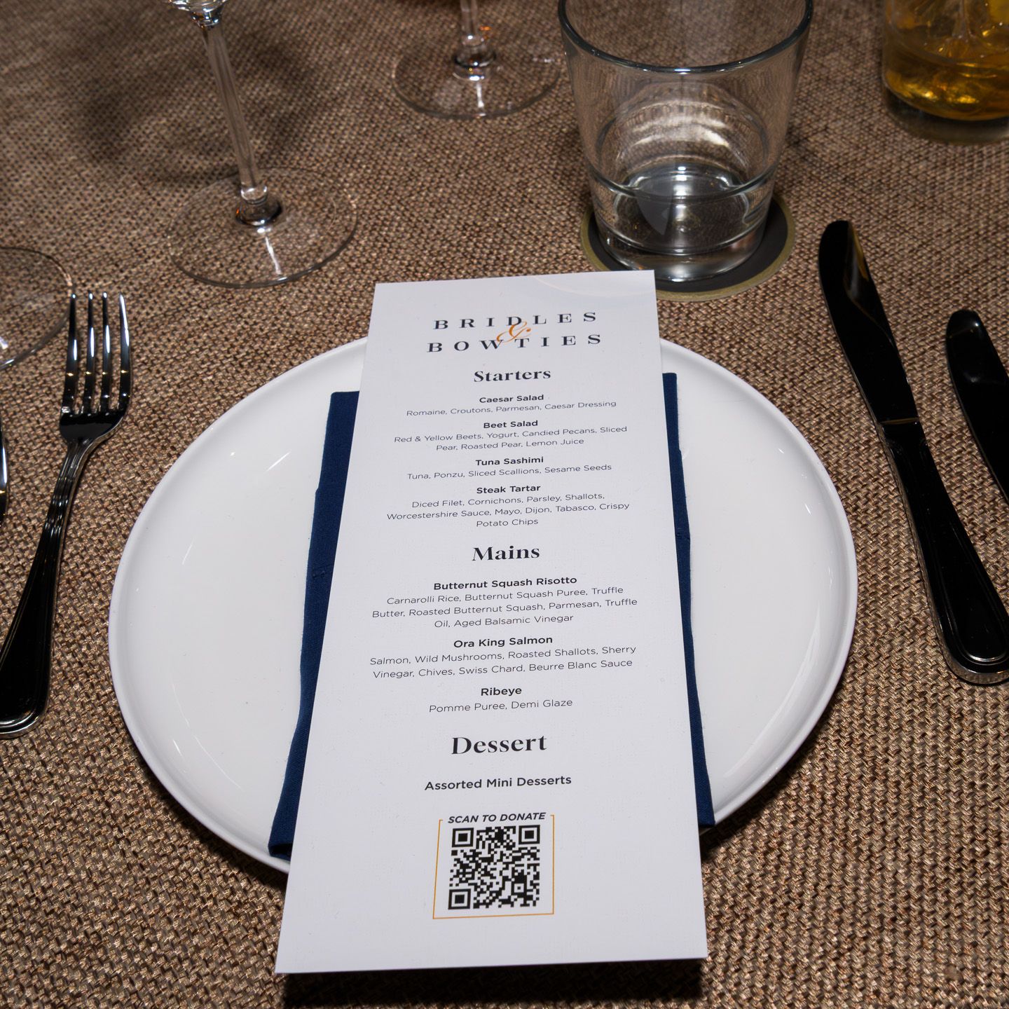 Elegant place setting with Bridles and Bowties event menu card, black cutlery, and wine glasses on burlap tablecloth.