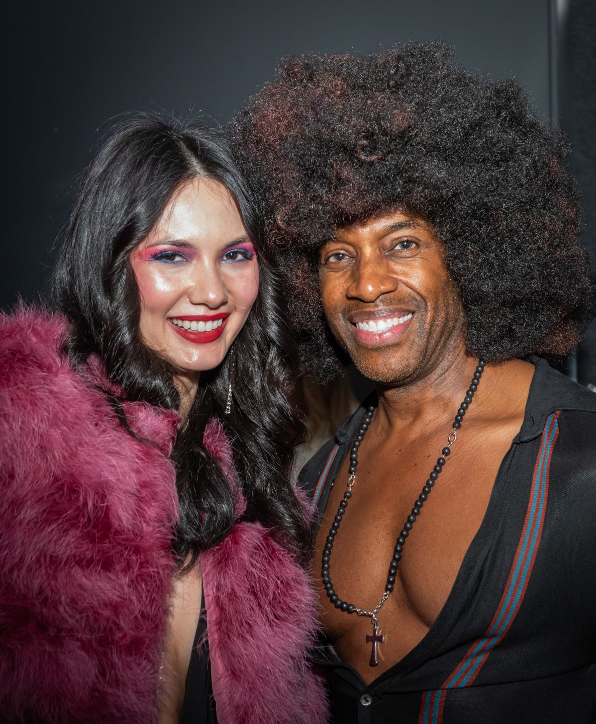 Smiling couple poses at a party, woman in pink fur coat and man with large afro hairstyle.