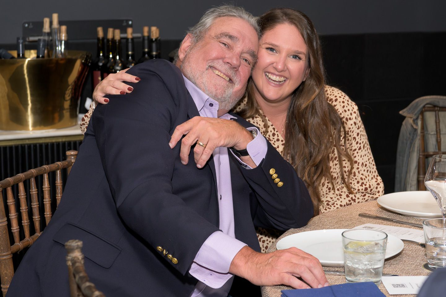 Happy older man and young woman share a warm embrace at a formal dinner.