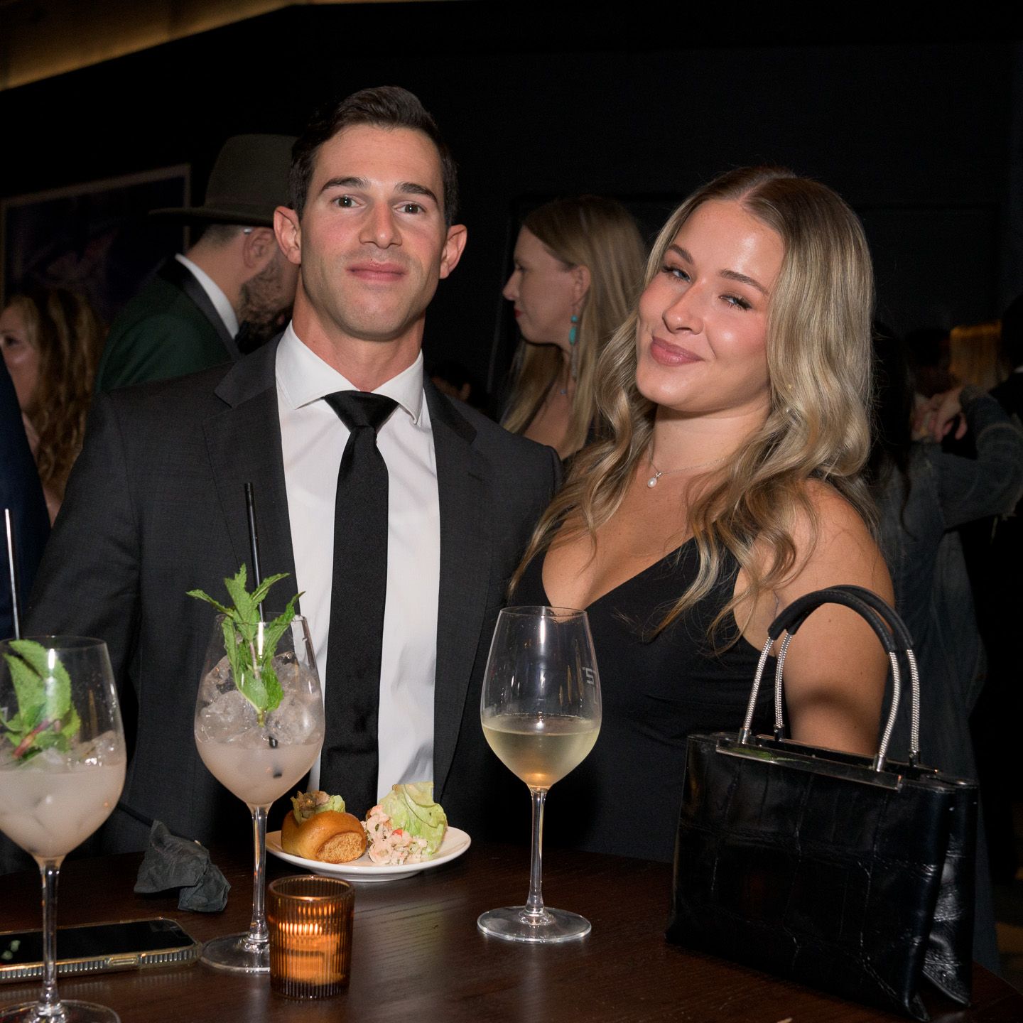 Couple smiling at bar with cocktails and wine glasses at evening event.