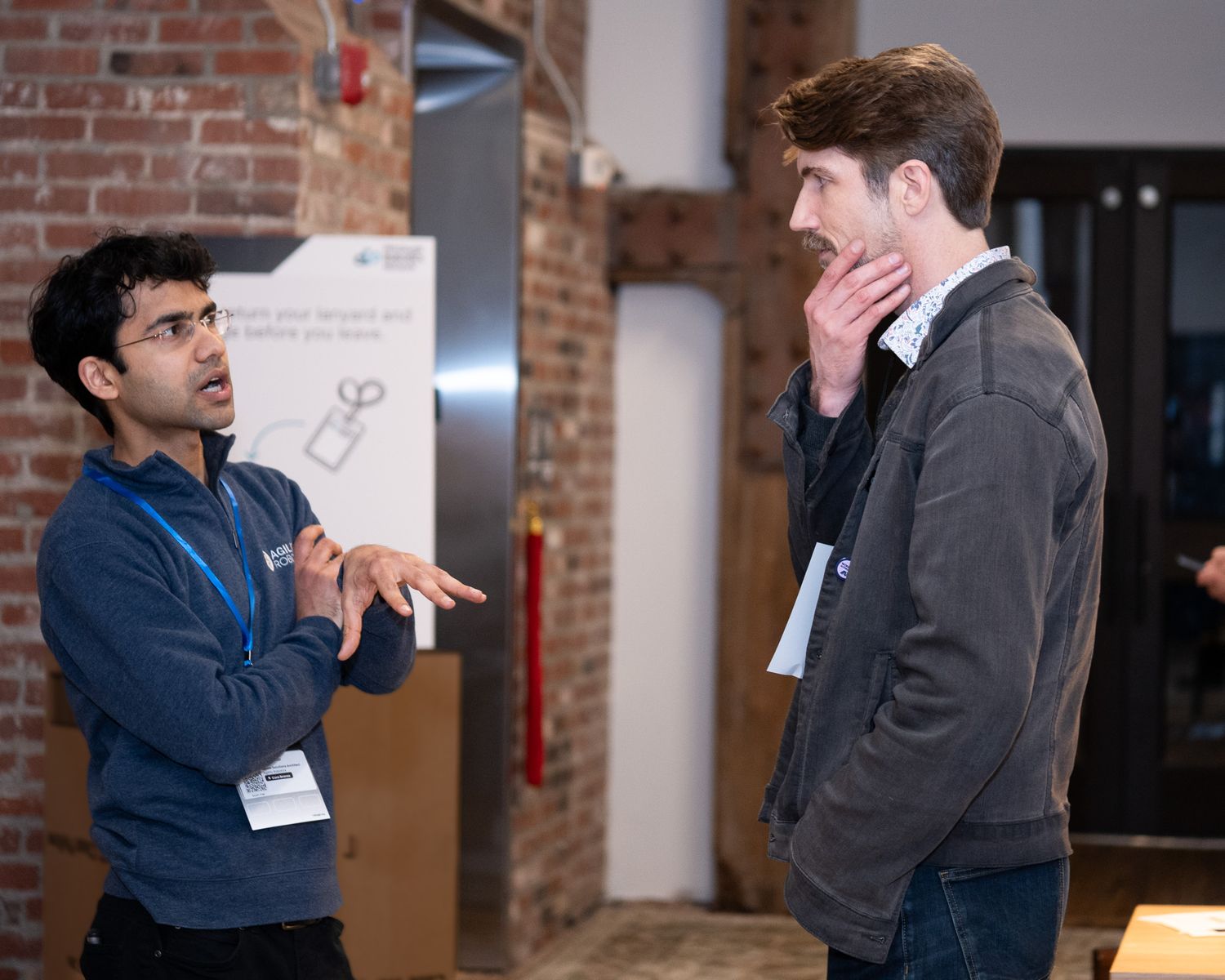 Two men engaged in conversation at an indoor networking event near a brick wall.