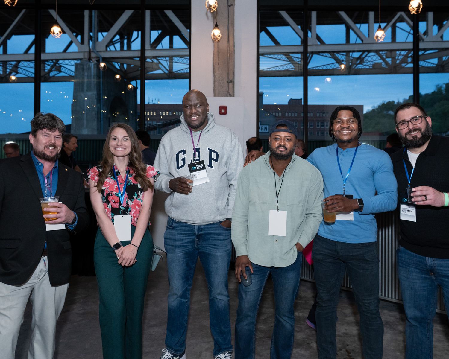 Group of six diverse professionals smiling together at an evening networking event near large windows.