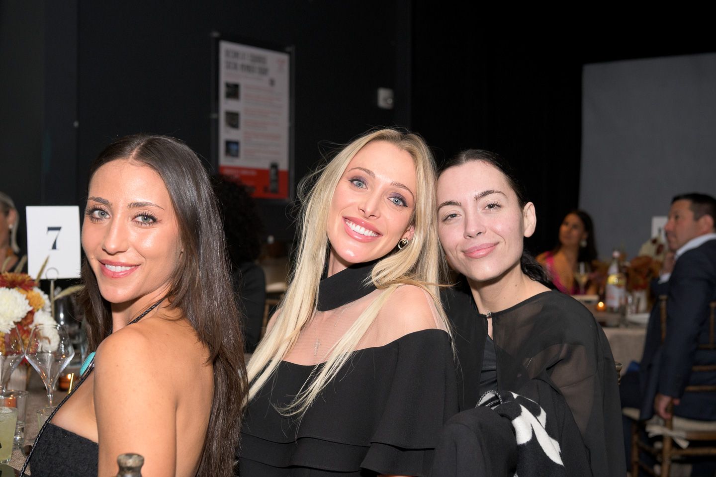Three women in black outfits smile together at a formal charity gala event.