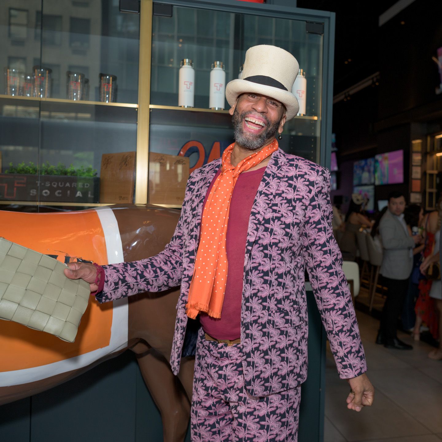 Stylish man in pink floral suit and white hat poses at an indoor venue event.