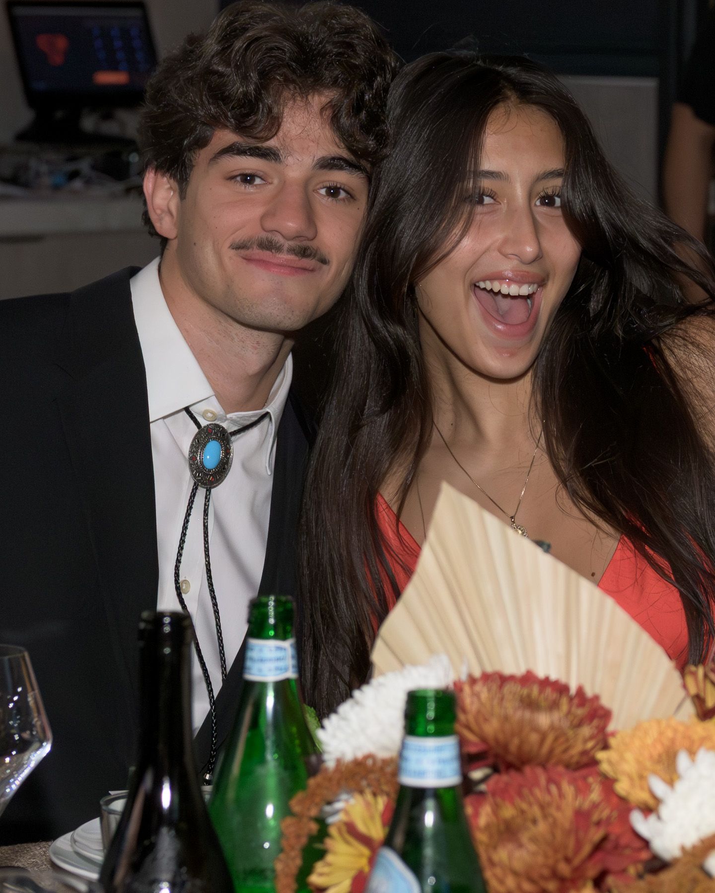 Young couple smiling at formal dinner event with wine bottles and floral centerpiece on table.