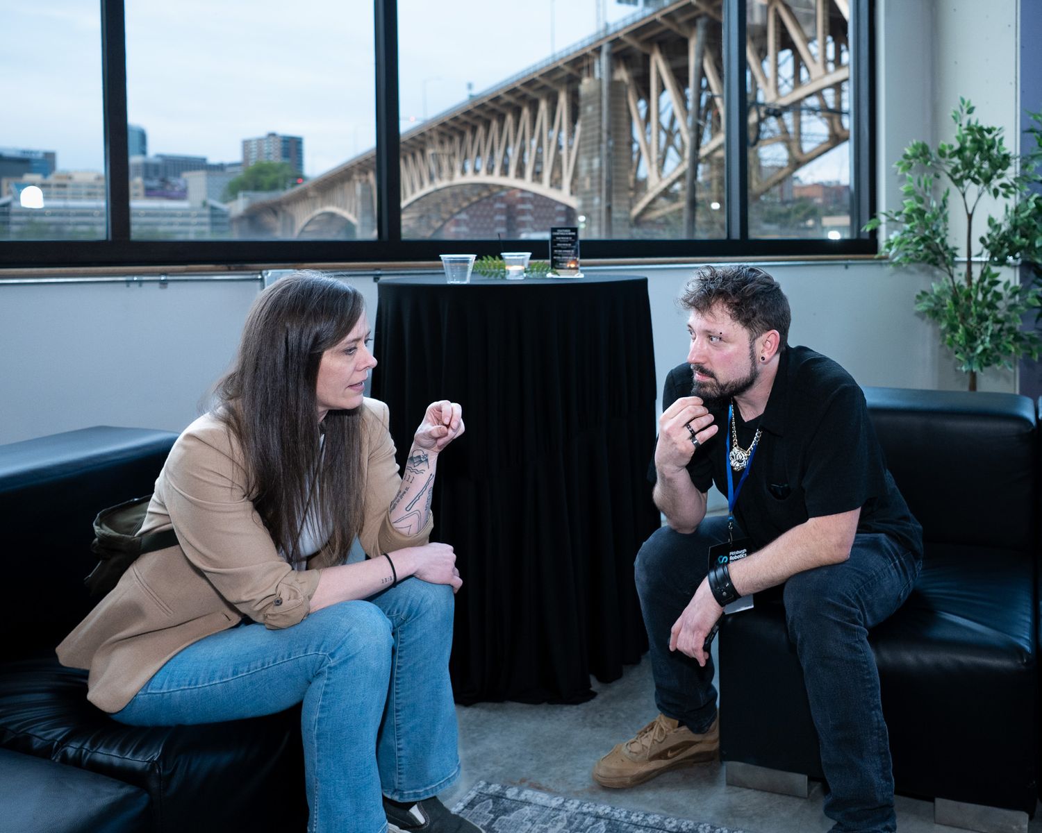 Man and woman having a conversation while seated on a black leather sofa at an indoor event.