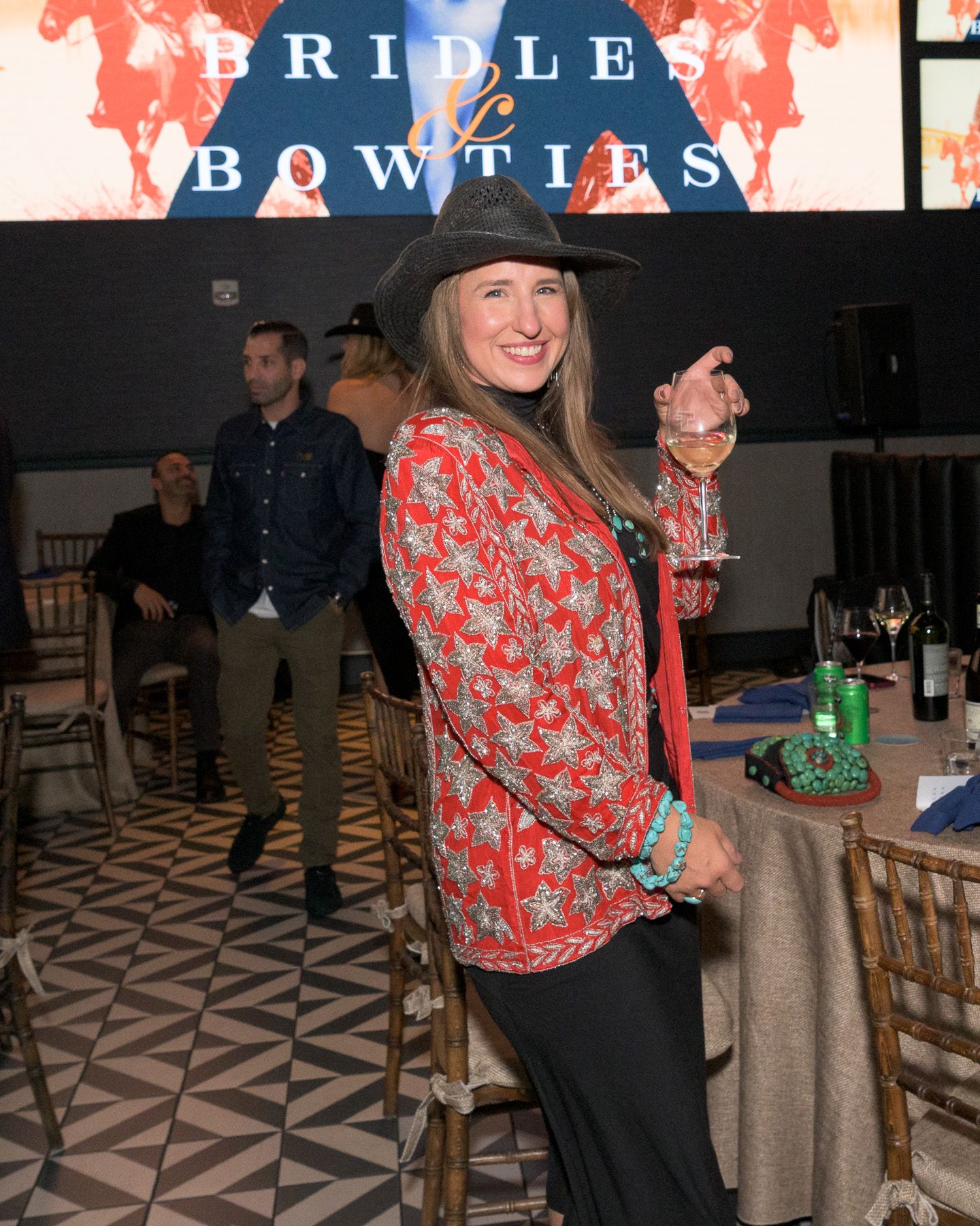 Woman in red floral jacket and black cowboy hat smiles at Bridles and Bowties charity gala event.